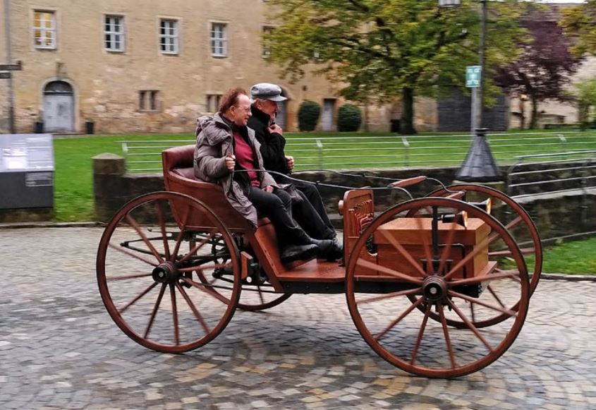Nachbau eines hölzernen Motorwagens von Andreas Flocken, Flocken Wagen genannt, während der Fahrt mit zwei Passagieren, 2025